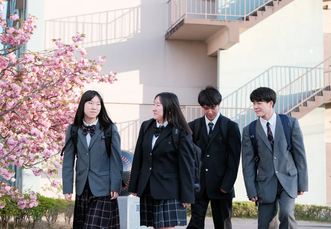校舎前で制服姿の学生たちが会話をしながら並んで歩く日常の登校風景