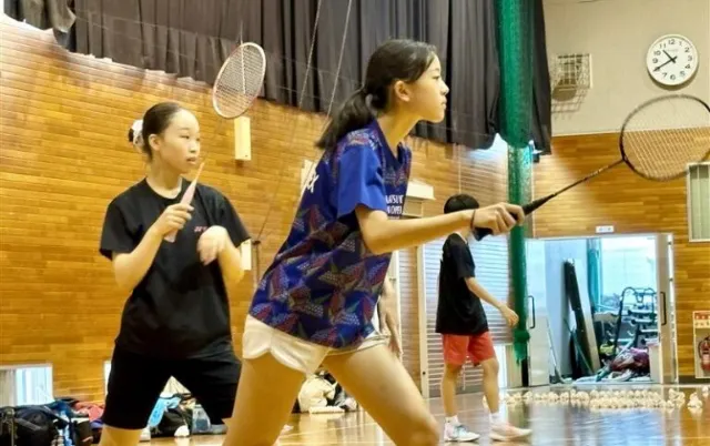 屋内の体育館で、バドミントン部の部員がラケットを構えている様子