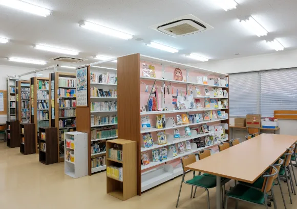 書籍が並ぶ書架と、閲覧用のテーブルと椅子が配置された図書室の室内